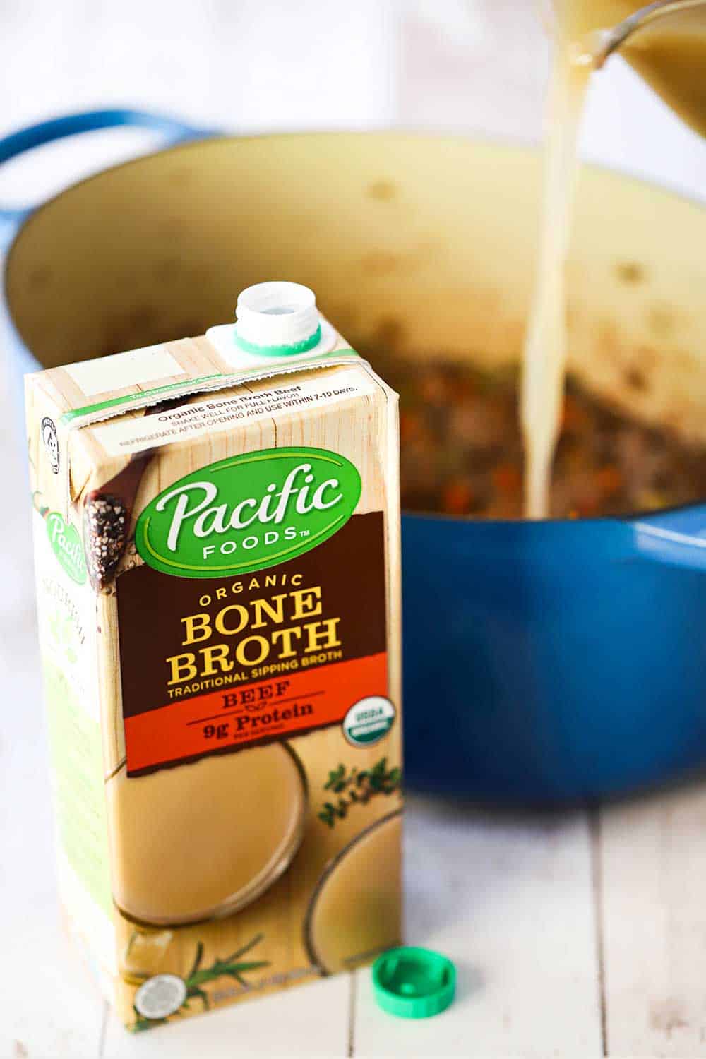 A box of Pacific Food bone broth sitting in front of a blue Dutch oven with a measuring cup pouring the broth into the pot.