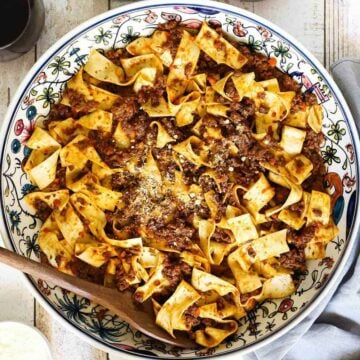 A large patterned Italian pasta bowl filled with Pappardelle Bolognese and a small bowl of Parmesan cheese next to it.