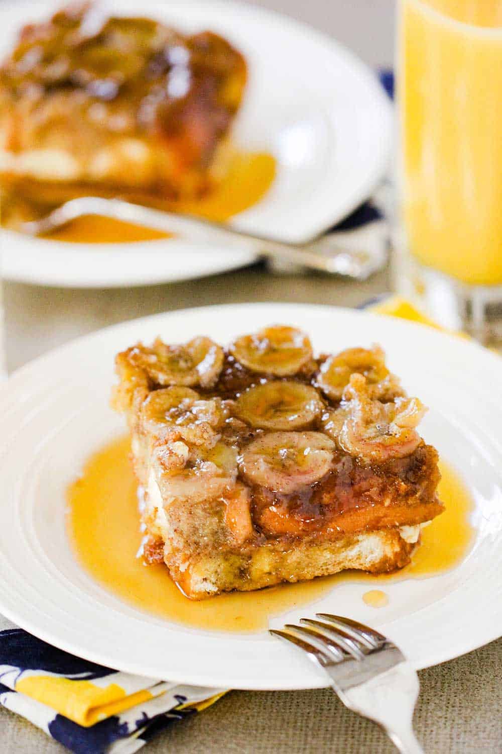 A piece of Caramel Banana Baked French Toast on a white plate with a fork on it and a glass of orange juice in the background.