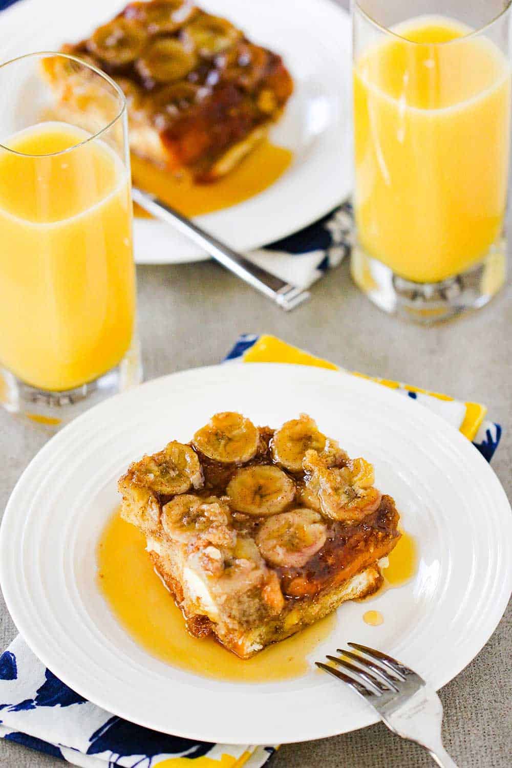 A view of two white plates each holding a slice of baked French toast with two glasses of orange juice next to them.