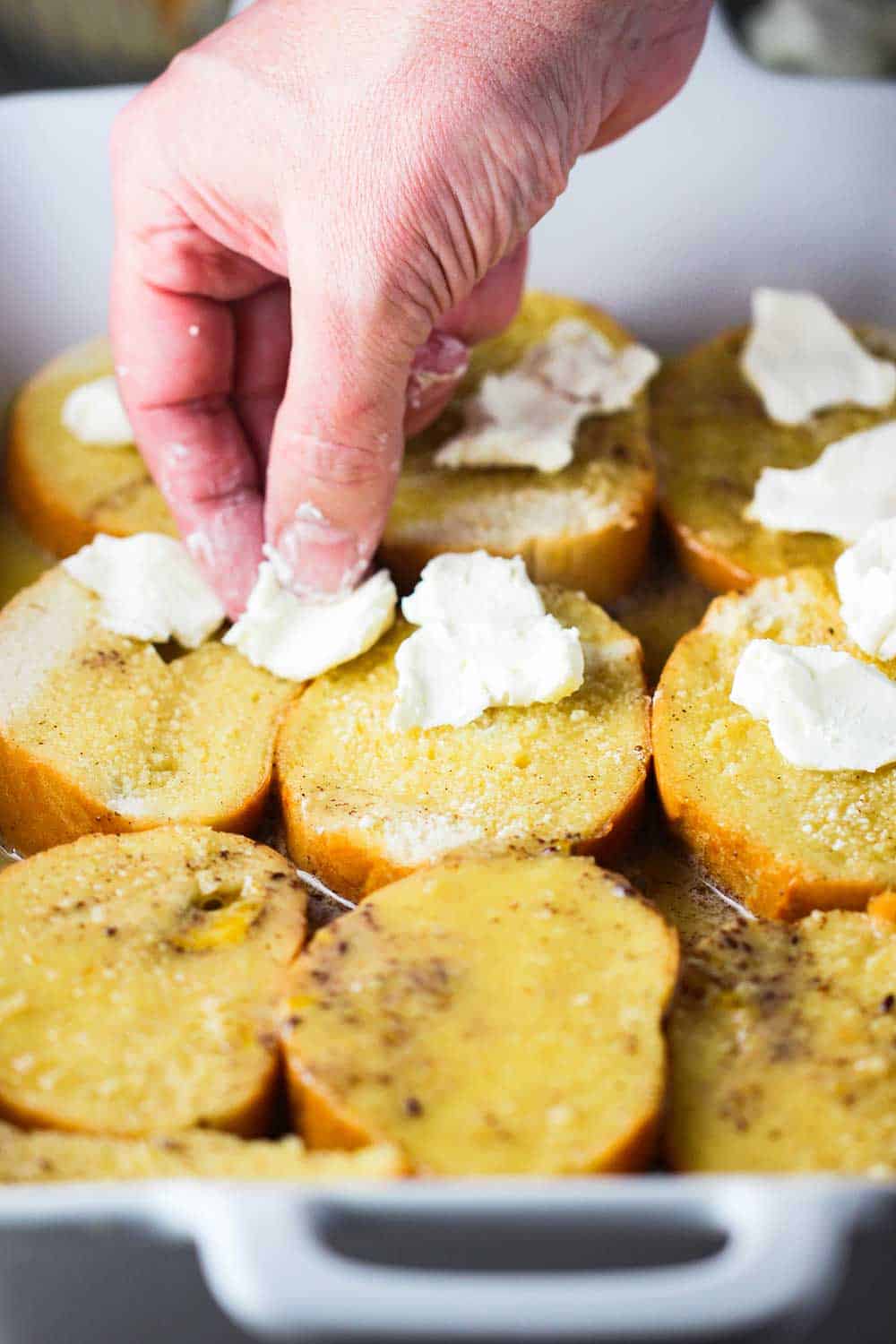 A hand placing pieces of cream cheese on top sliced bread for baked French toast.