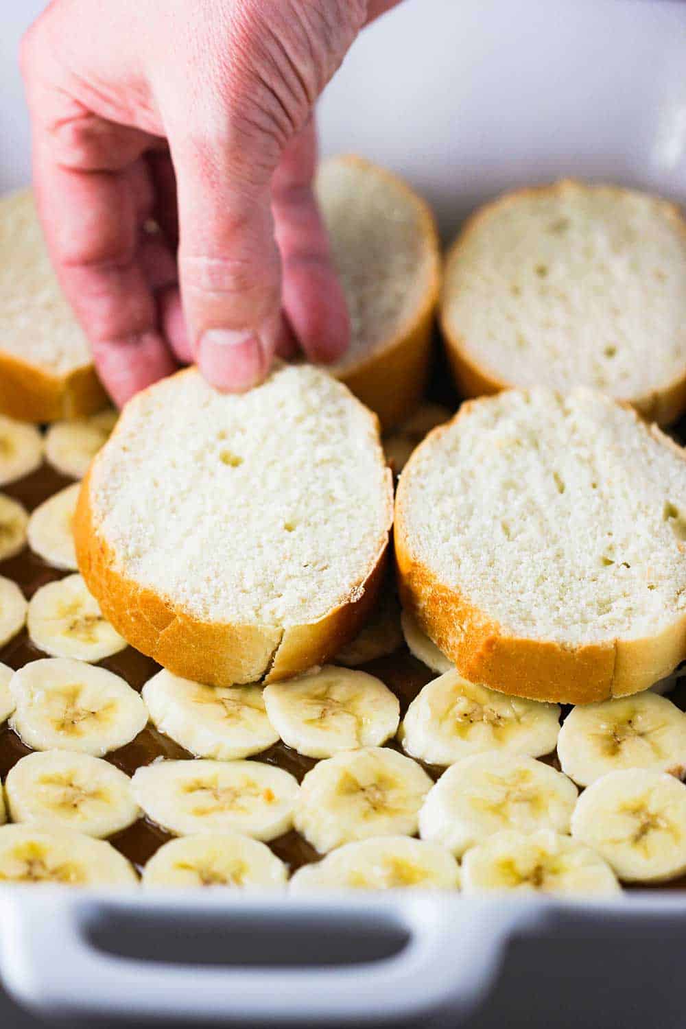 A hand placing slices of French bread over a layer of slice bananas and caramel sauce in a white baking dish.