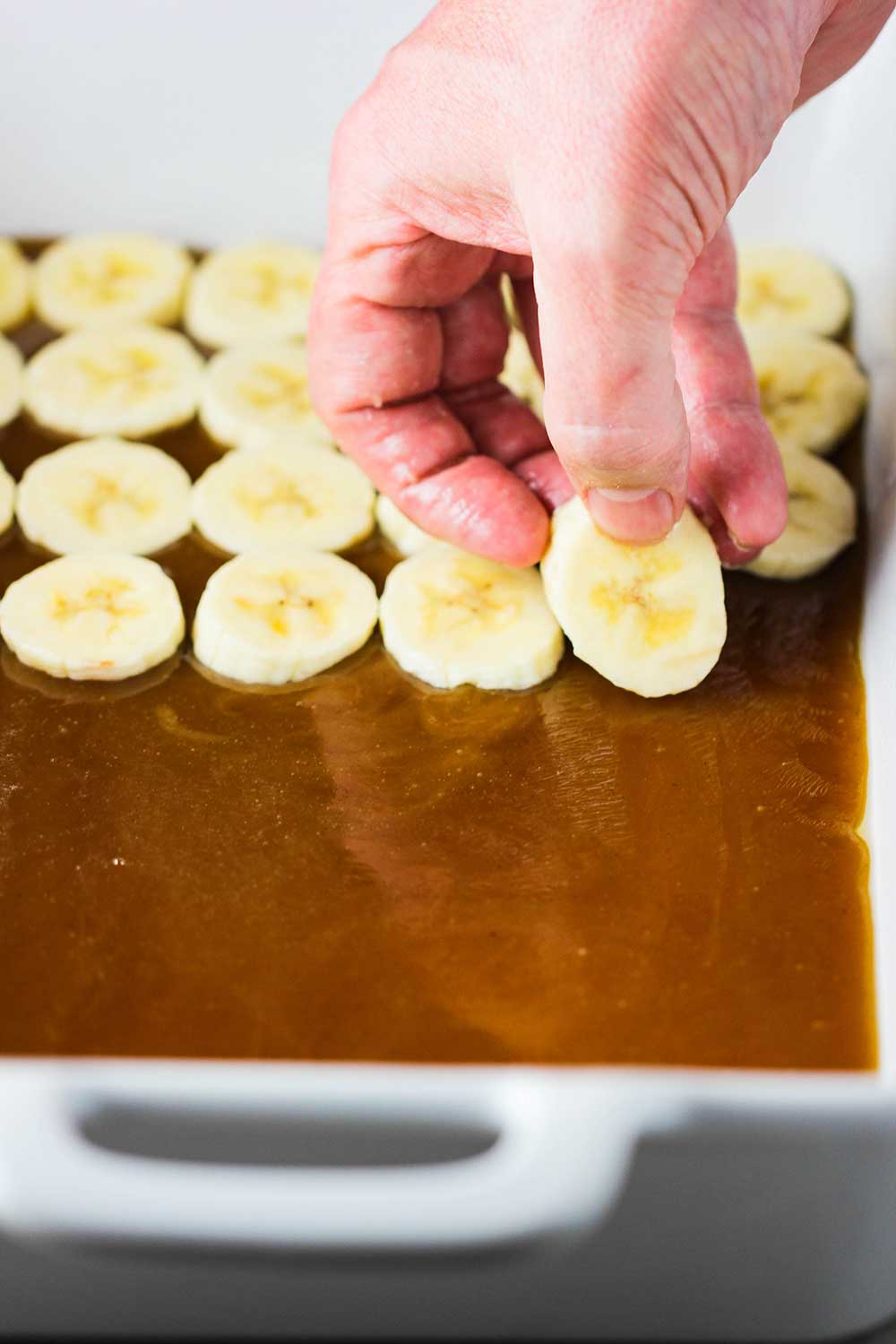 A hand placing sliced bananas on top of a layer of caramel sauce in a white baking dish.