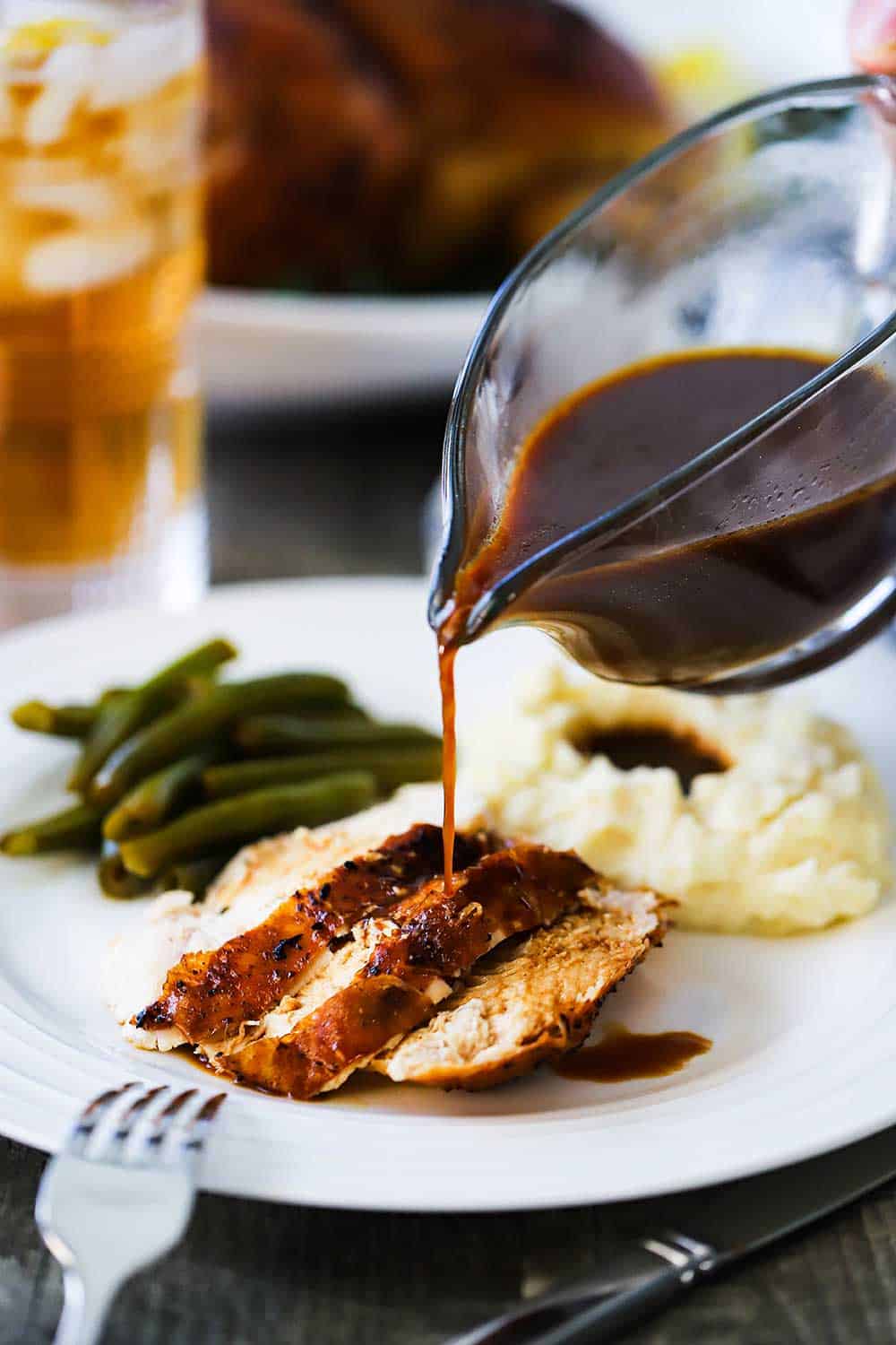 Brown gravy being poured from a glass gravy boat over sliced roasted chicken next to mashed potatoes and green beans.