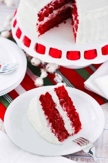 A slice of red velvet cake on a white plate next to the cake on a cake stand with red ribbon.