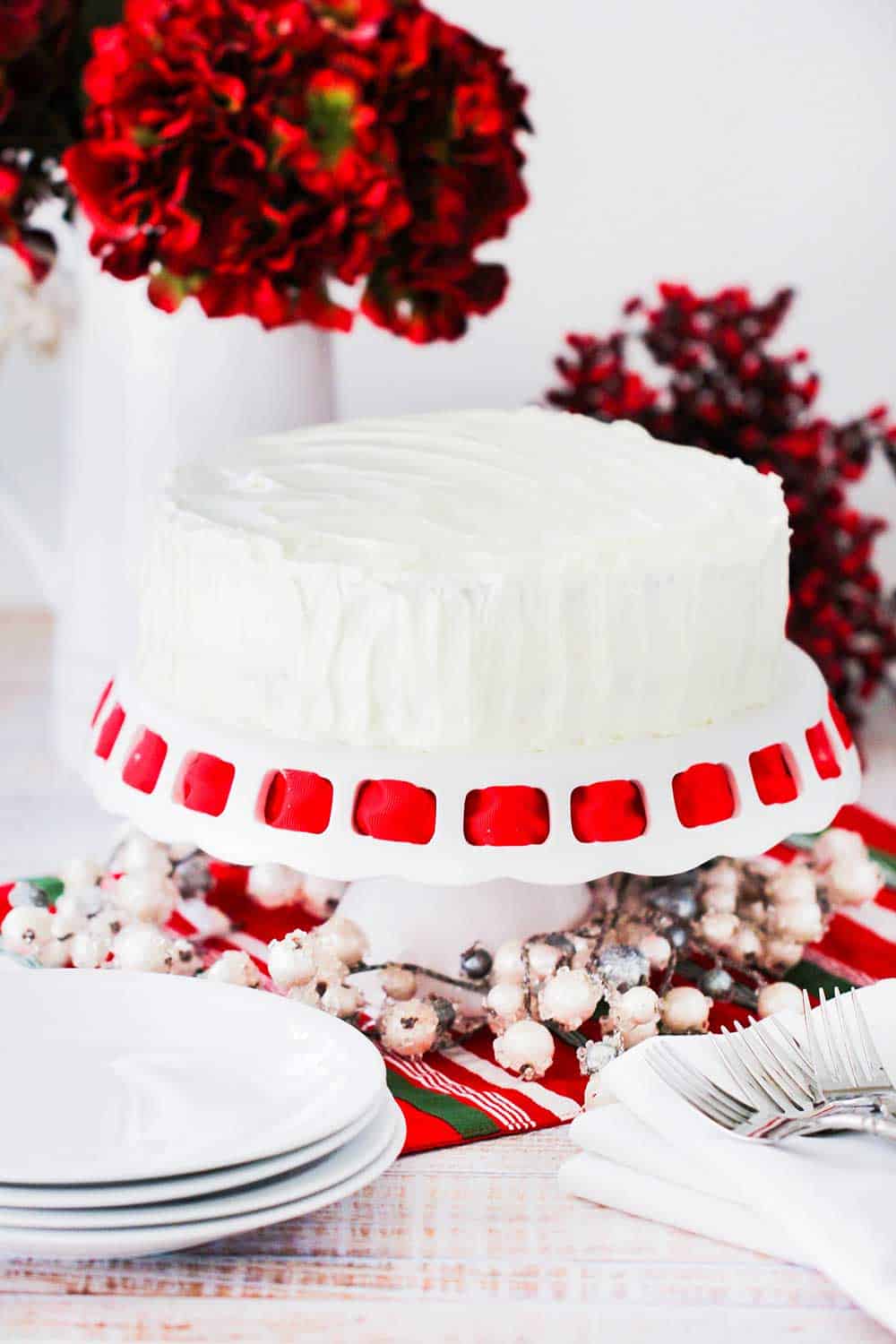 A red velvet cake on a cake stand next to a stack of white plates and a bouquet of red flowers nearby.