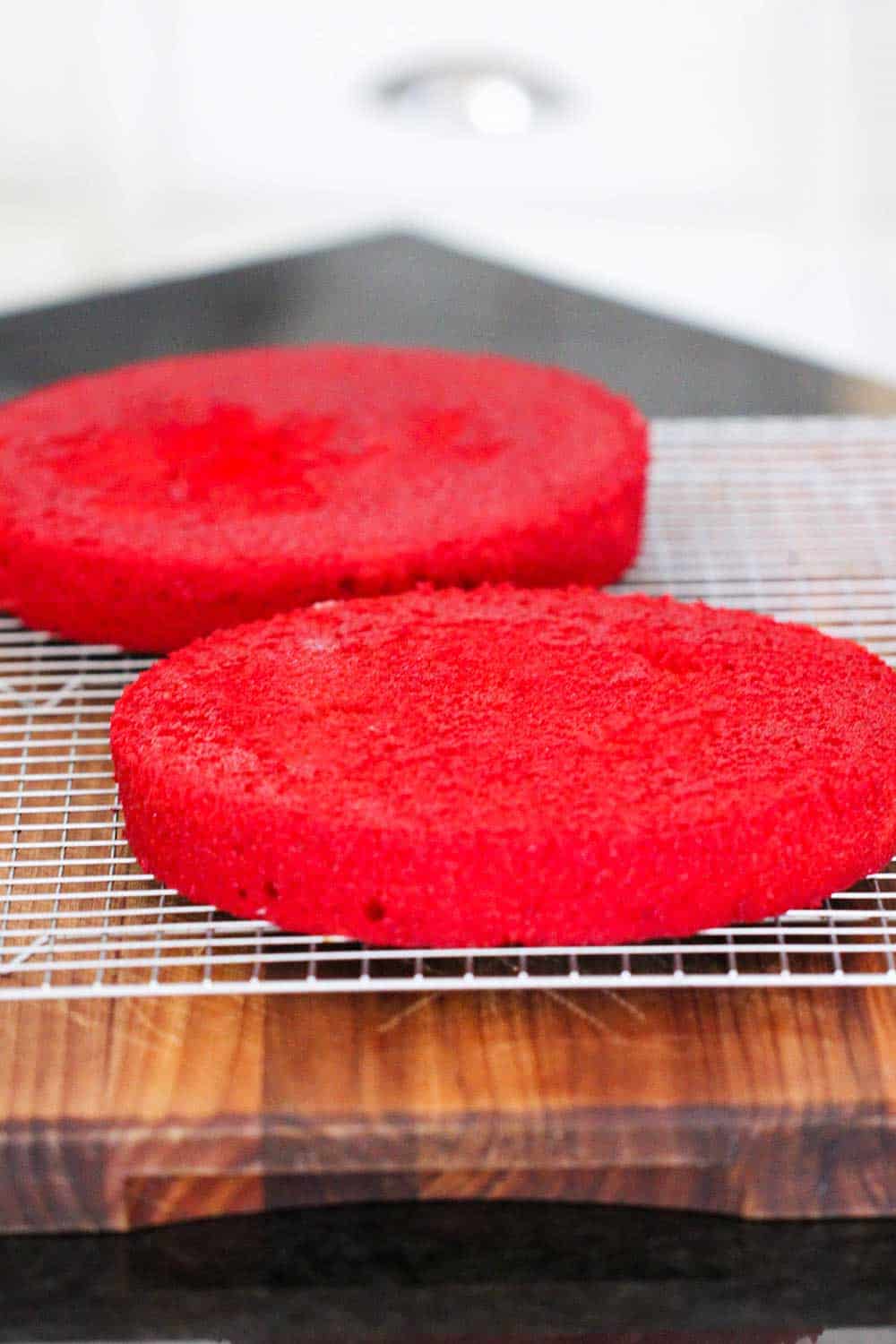 Two cooked layers of red velvet cake on a baking rack on a wooden cutting board.