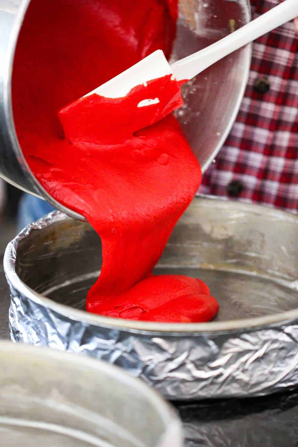 Red cake batter being poured into a cake pan lined with foil.