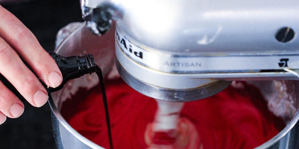 A hand pouring red food dye into batter in a stand mixer for red velvet cake.