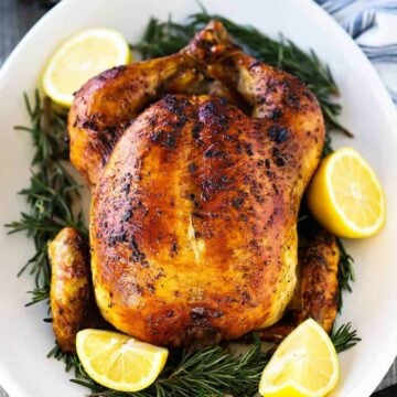 A roast chicken sitting on an oval platter with sliced lemons and herbs surrounding the bird.