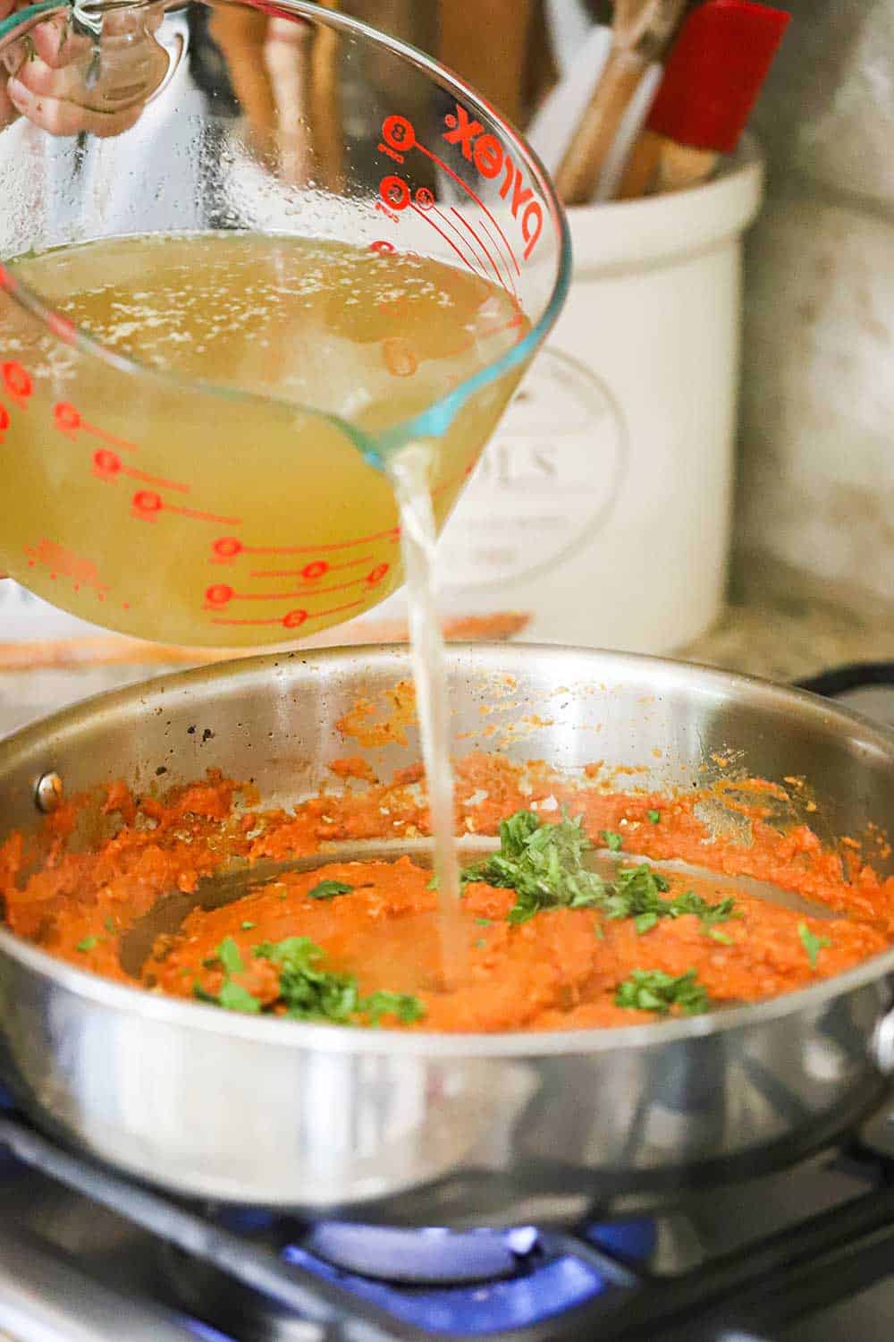 A large measuring cup of chicken broth being poured into a skillet with tomato paste and chopped cilantro.