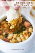 A hand dipping a torn piece of bread dipping into a bowl of smoked sausage and white bean stew.