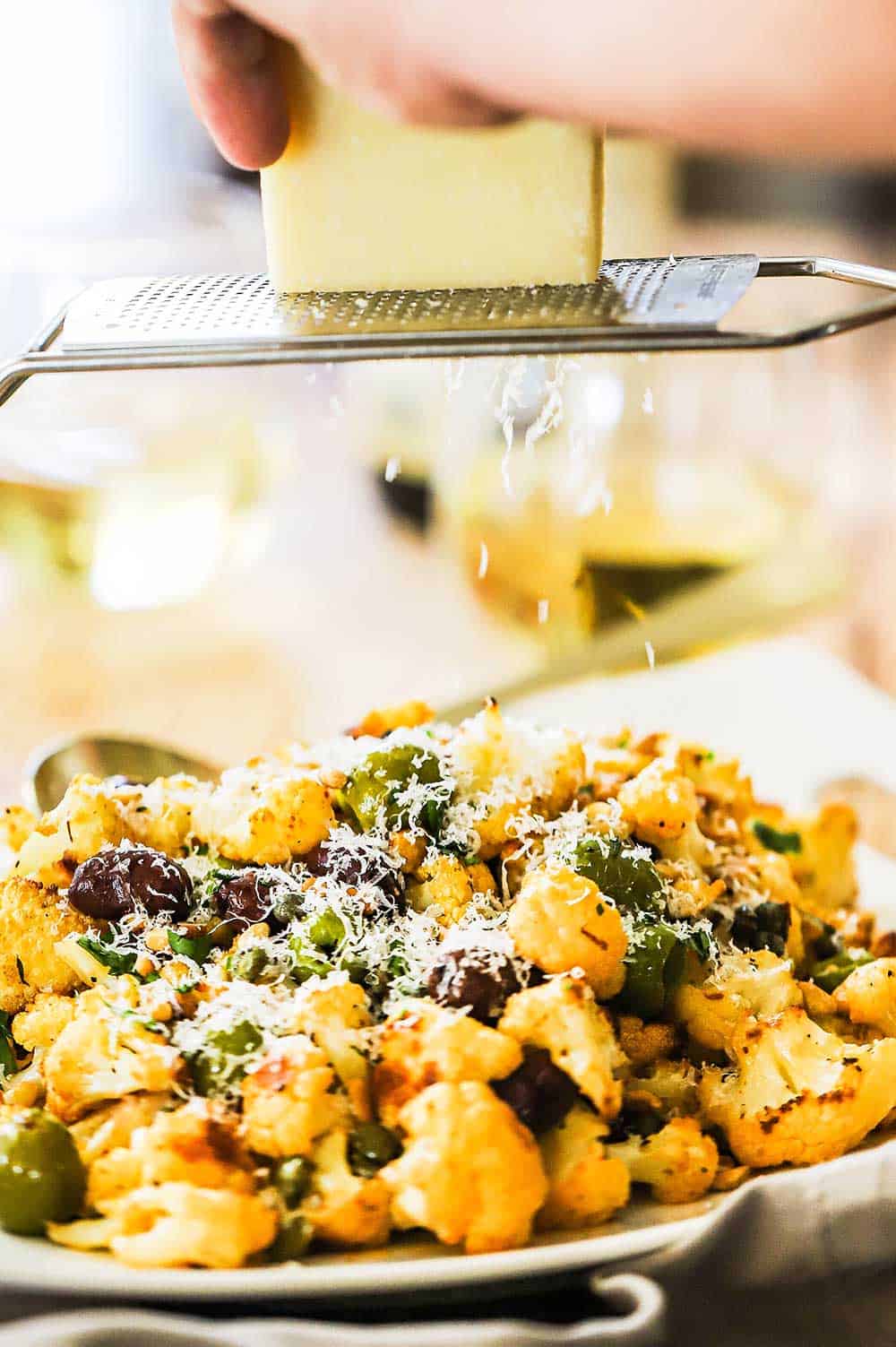 A person using a micro-plane to grate a block of Parmesan cheese over a platter of roasted cauliflower and olives.
