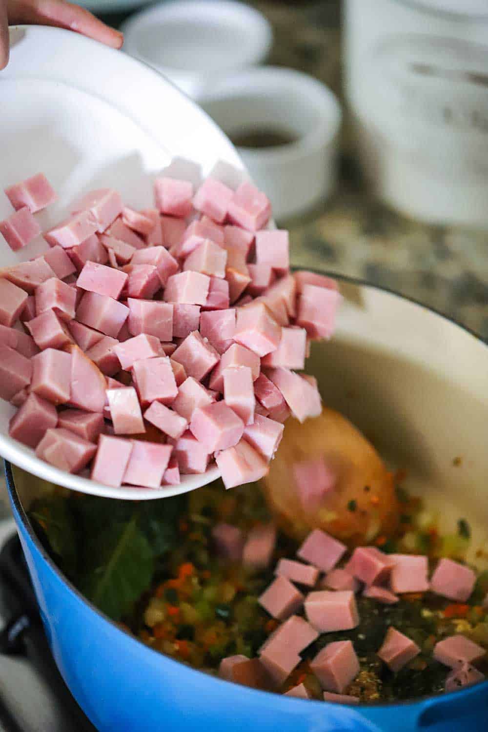 A bowl of cubed smoked ham being dropped into a Dutch oven filled with sautéed vegetables.