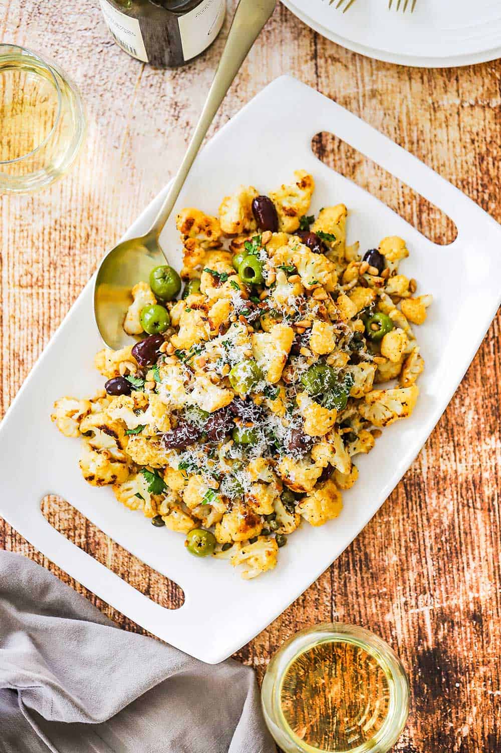 A white rectangular serving platter filled with roasted cauliflower with olives garnished with parsley and grated Parmesan.
