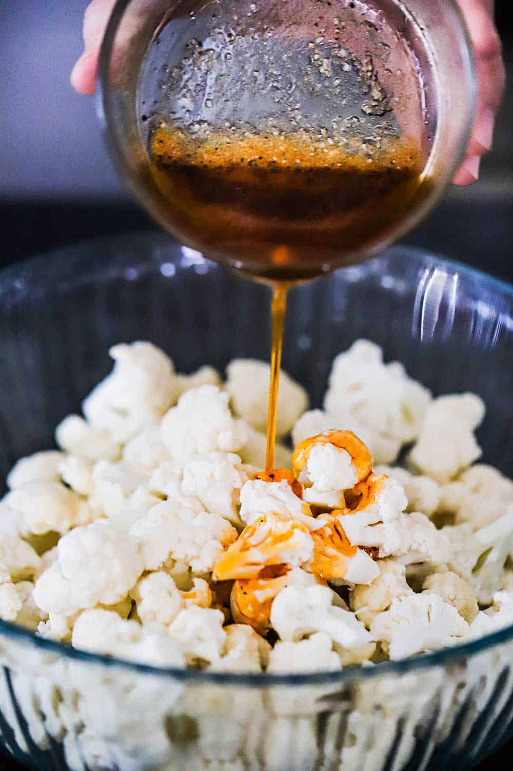 A person pouring paprika oil over cauliflower florets in a large glass bowl.