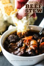 A hand dunking a torn piece of white bread into a bowl of hearty beef stew.