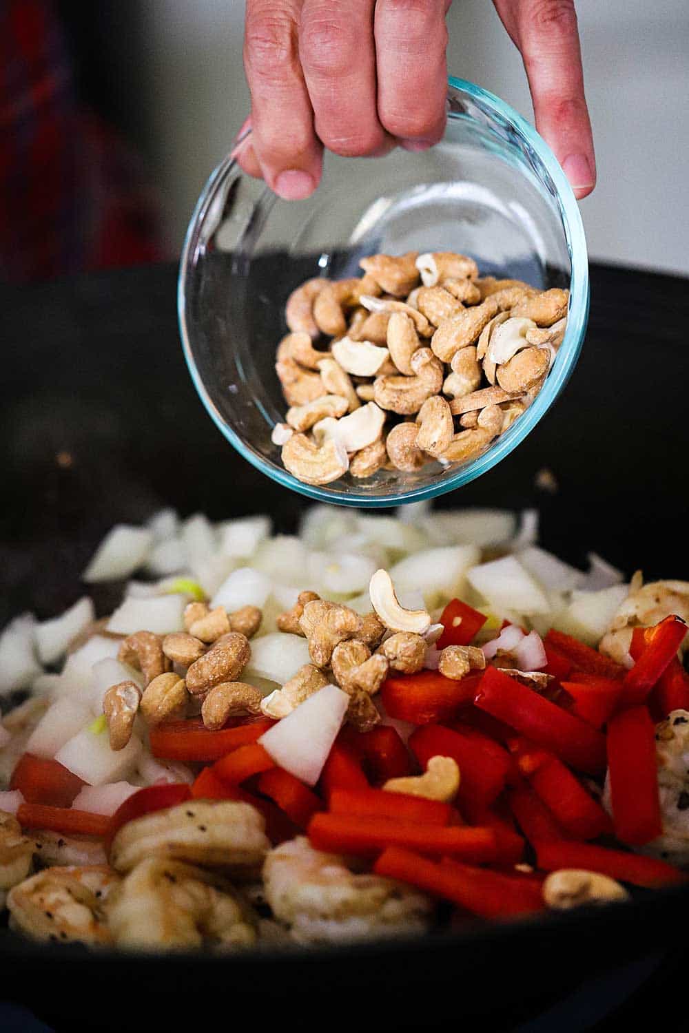 A person dumping whole roasted cashews from a small glass bowl into a wok filled with cooked shrimp and chopped onion and red bell pepper.