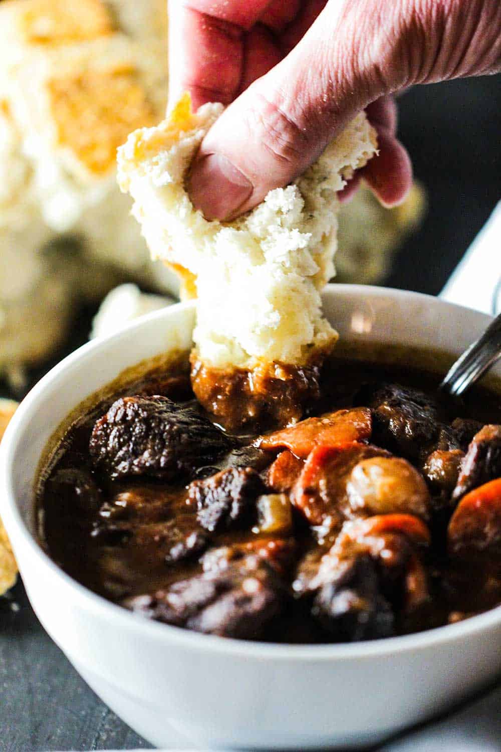 A hand dunking a torn piece of white bread into a bowl of hearty beef stew.