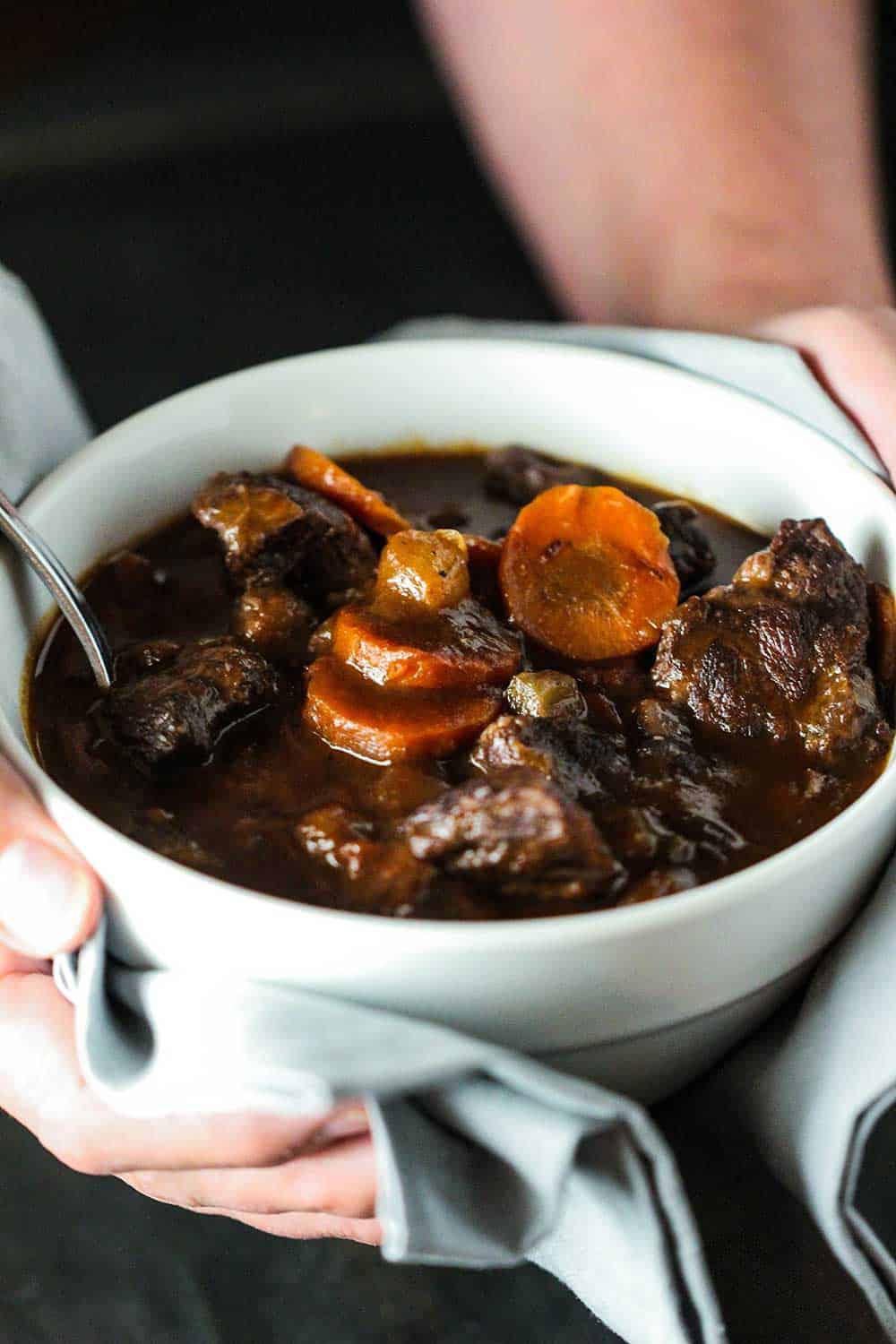 Two hands holding a white bowl filled with hearty beef stew.