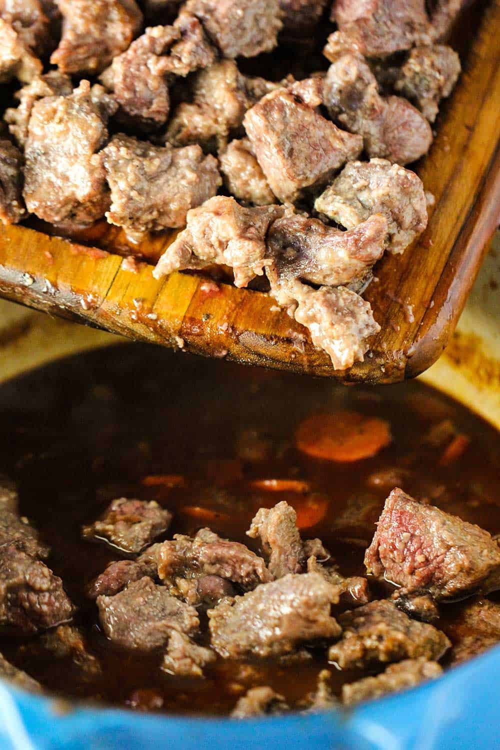 Seared beef being dropped into hearty beef stew from a cutting board.