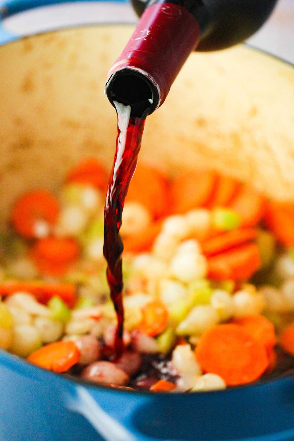 A bottle of red wine being poured into a blue Dutch oven filled with sautéed vegetables.