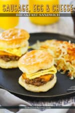 Two sausage, egg, and cheese breakfast sandwiches on a dark plate with hashbrowns in front a glass of orange juice.