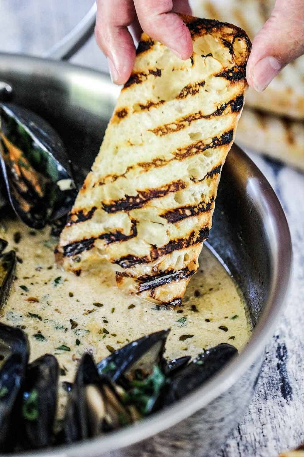 A hand dipping grilled bread into a pan of mussels with garlic and wine sauce.