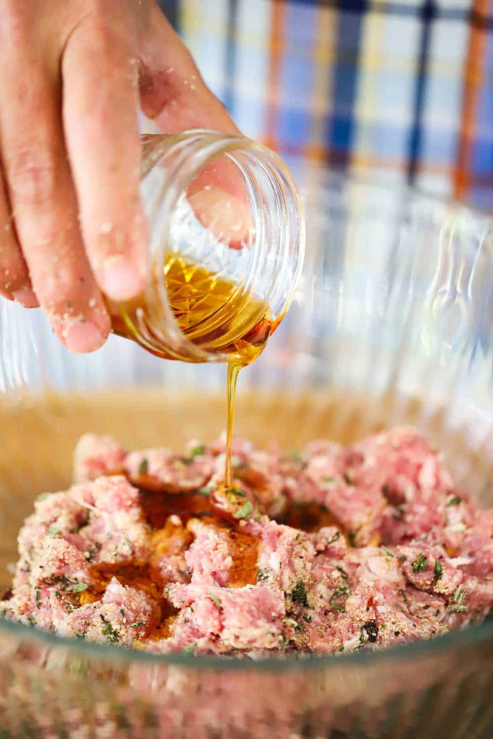 A hand pouring maple syrup from a small jar into a bowl of seasoned ground pork.