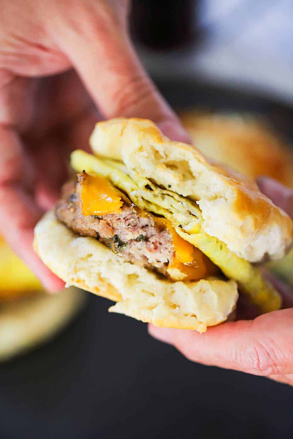 Two hands holding a sausage, egg, and cheese breakfast sandwich with a bite taken out it.