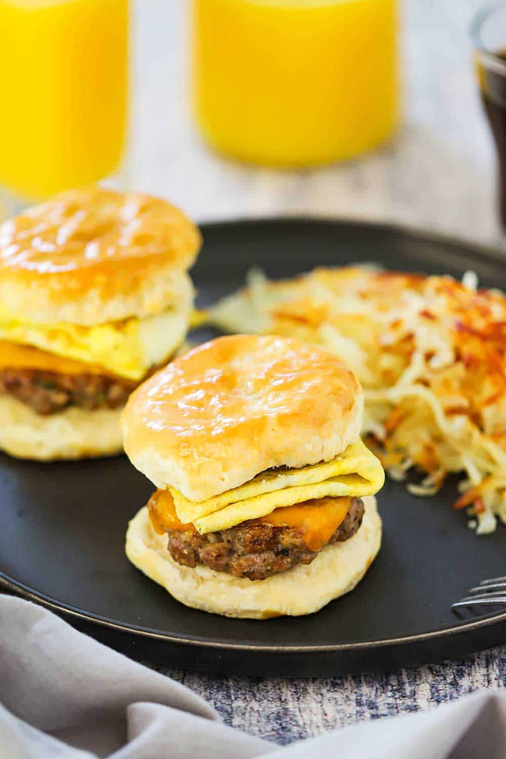 Two sausage, egg, and cheese breakfast sandwiches on a dark plate with hashbrowns in front a glass of orange juice.