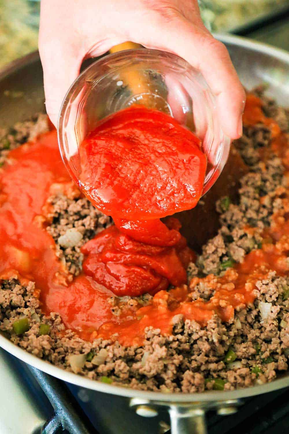 A hand pouring a small glass bowl of chili sauce into a skillet of cooked ground beef.