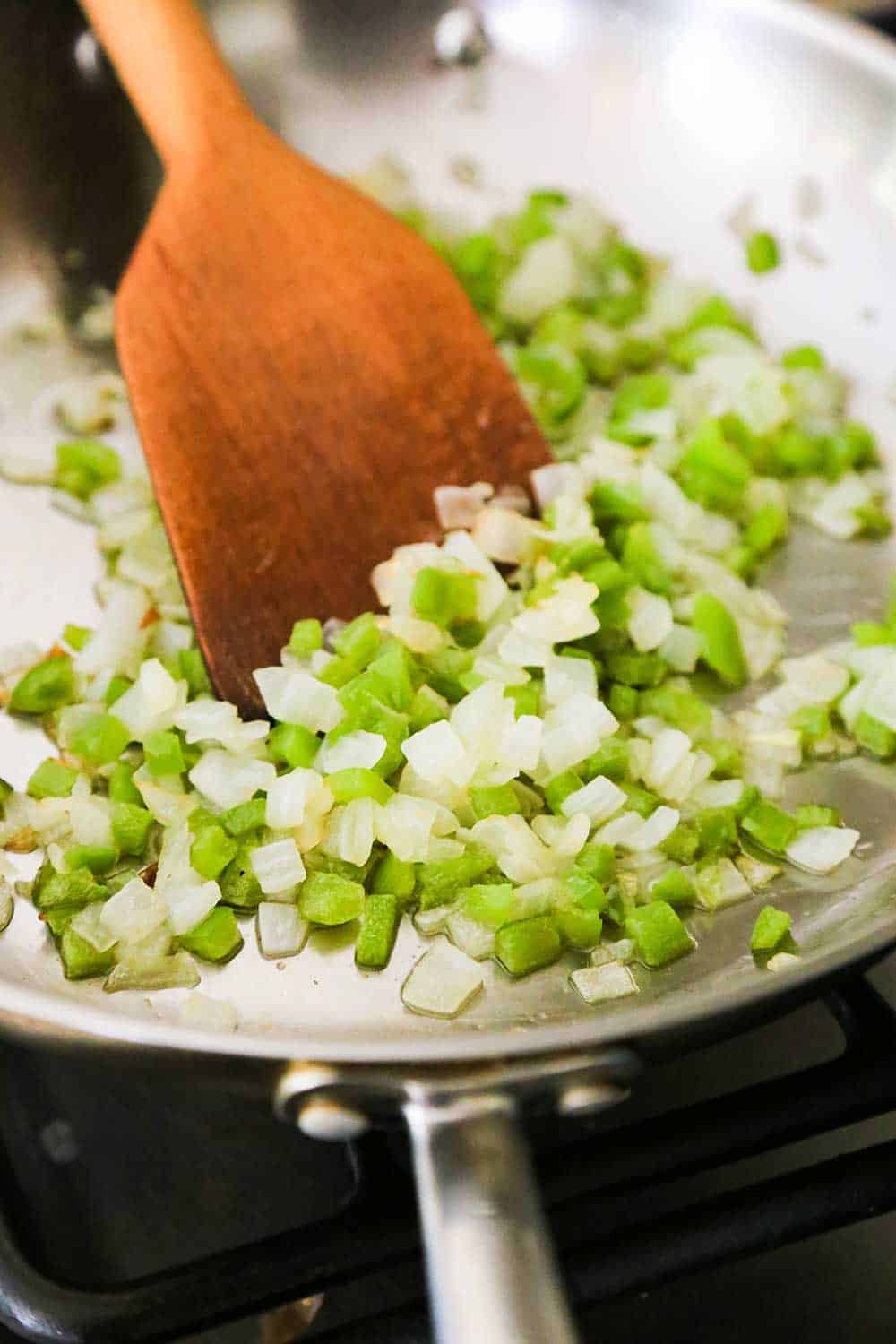 A large silver skillet filled with sautéed green bell peppers and onion with a wooden spoon in it.