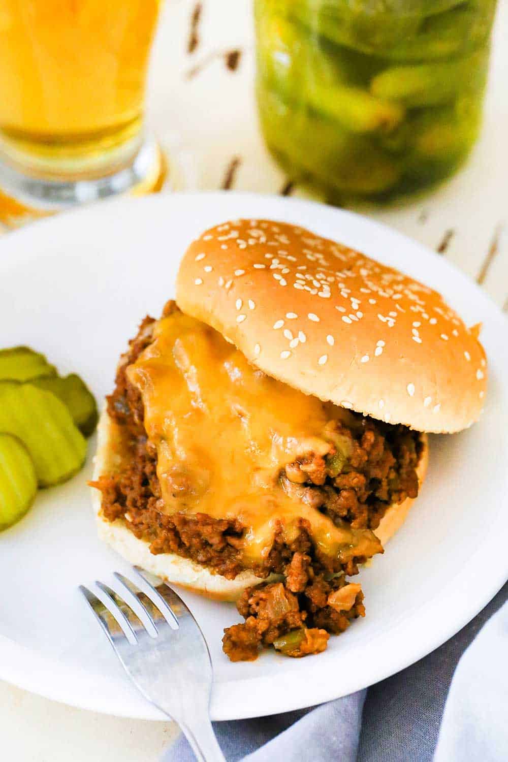 A sloppy joes sandwich on a white plate next to a jar of sliced pickles.