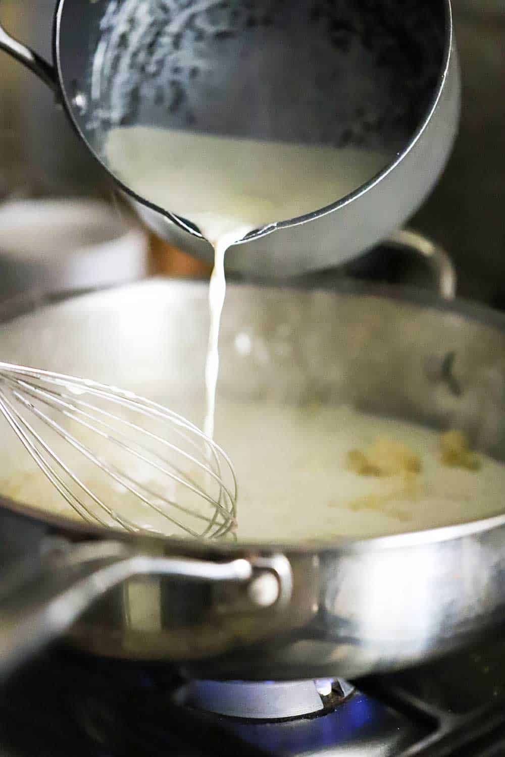 A medium-sized saucepan filled with cream is being poured into a skillet with a roux in it.