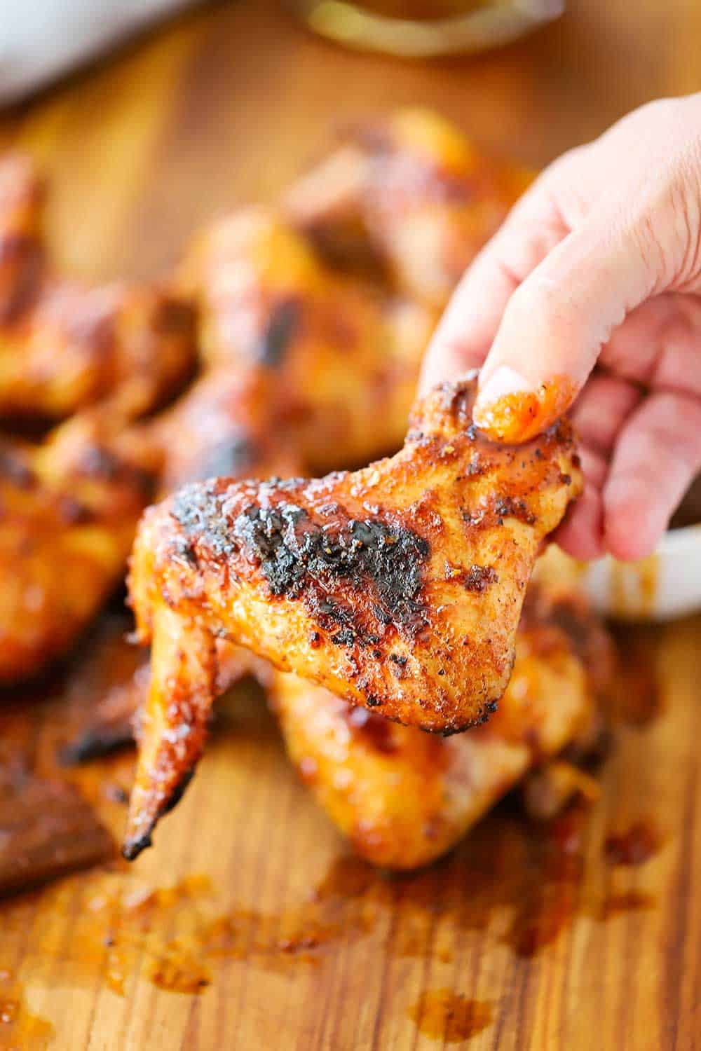 A hand holding up a grilled bourbon maple wing over a wooden cutting board filled with the same.