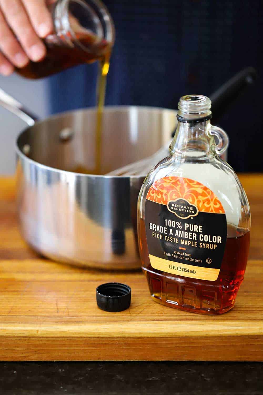 A hand pouring a jar of pure maple syrup into a stainless steel saucepan next to a bottle of the pure maple syrup all on a wooden cutting board.
