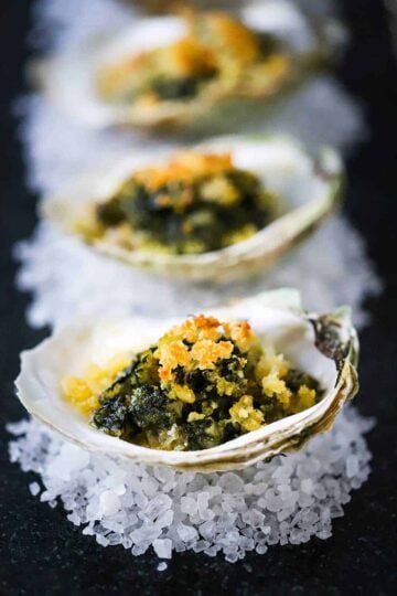 A row of oysters Rockefeller each sitting on rock salt on a dark grey serving tray.