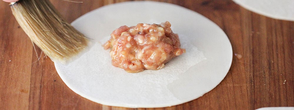 A brush applying water along the edges of a dumpling wrapper for pork dumplings with spicy peanut sauce.