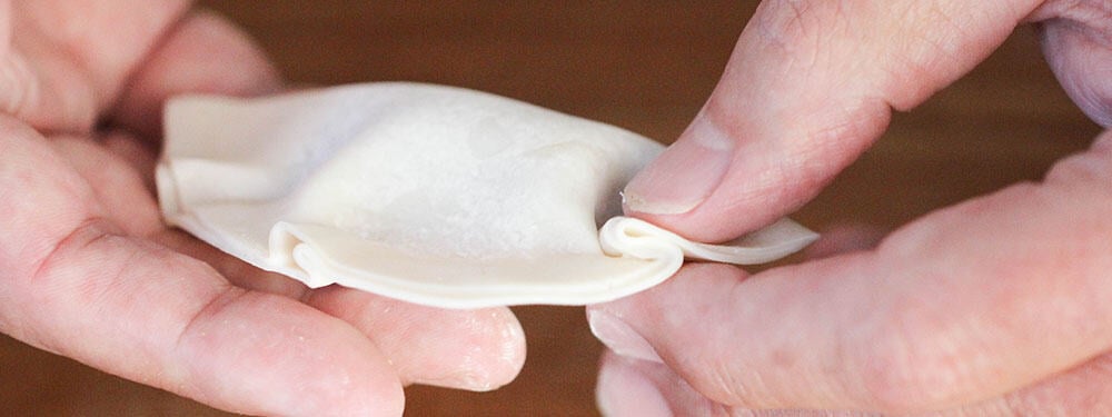 Two thumbs creating a pleat in a pork dumpling wrapper.