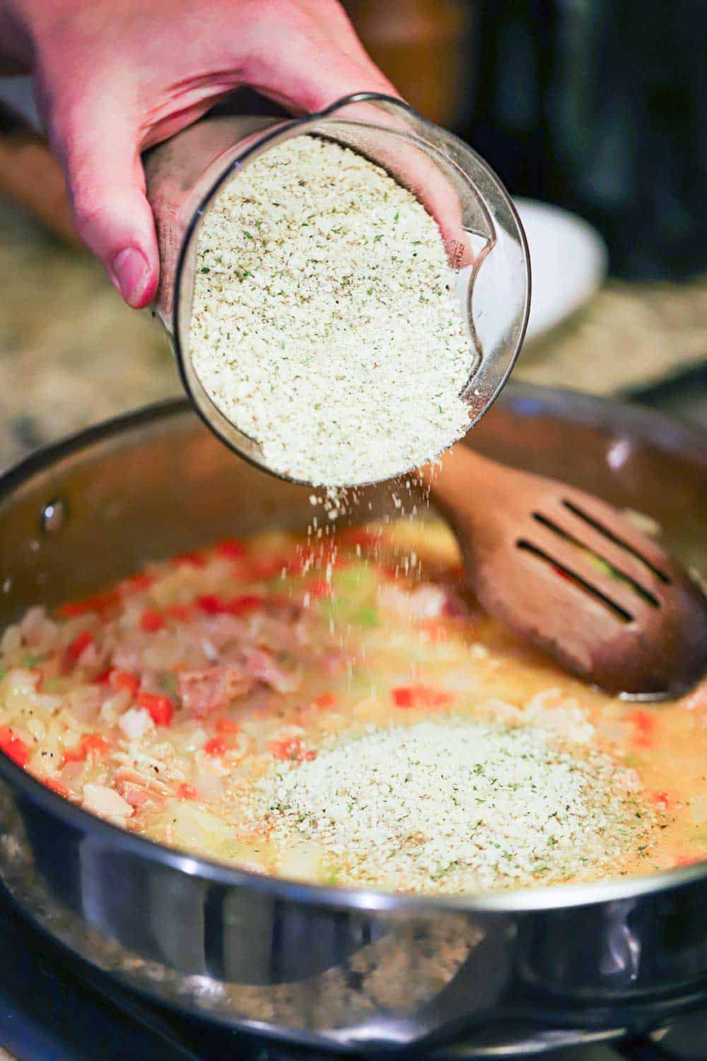 A hand dumping Italian-style Panko breadcrumbs into a skillet filled with clams, clam juice, and sautéd vegetables.