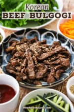 A close-up view of a shallow blue bowl that is filled with cooked bulgogi beef strips and surrounded by other small bowls of pickled vegetables and lettuce leaves.
