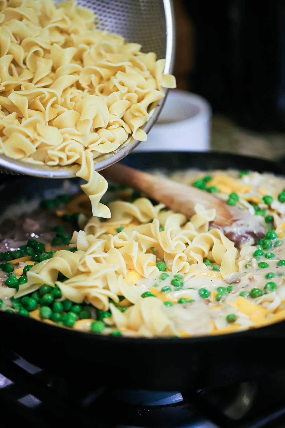 Cooked extra-wide egg noodles being transferred into a large cast-iron skillet filled with cream of mushroom soup, peas, and shredded cheese.
