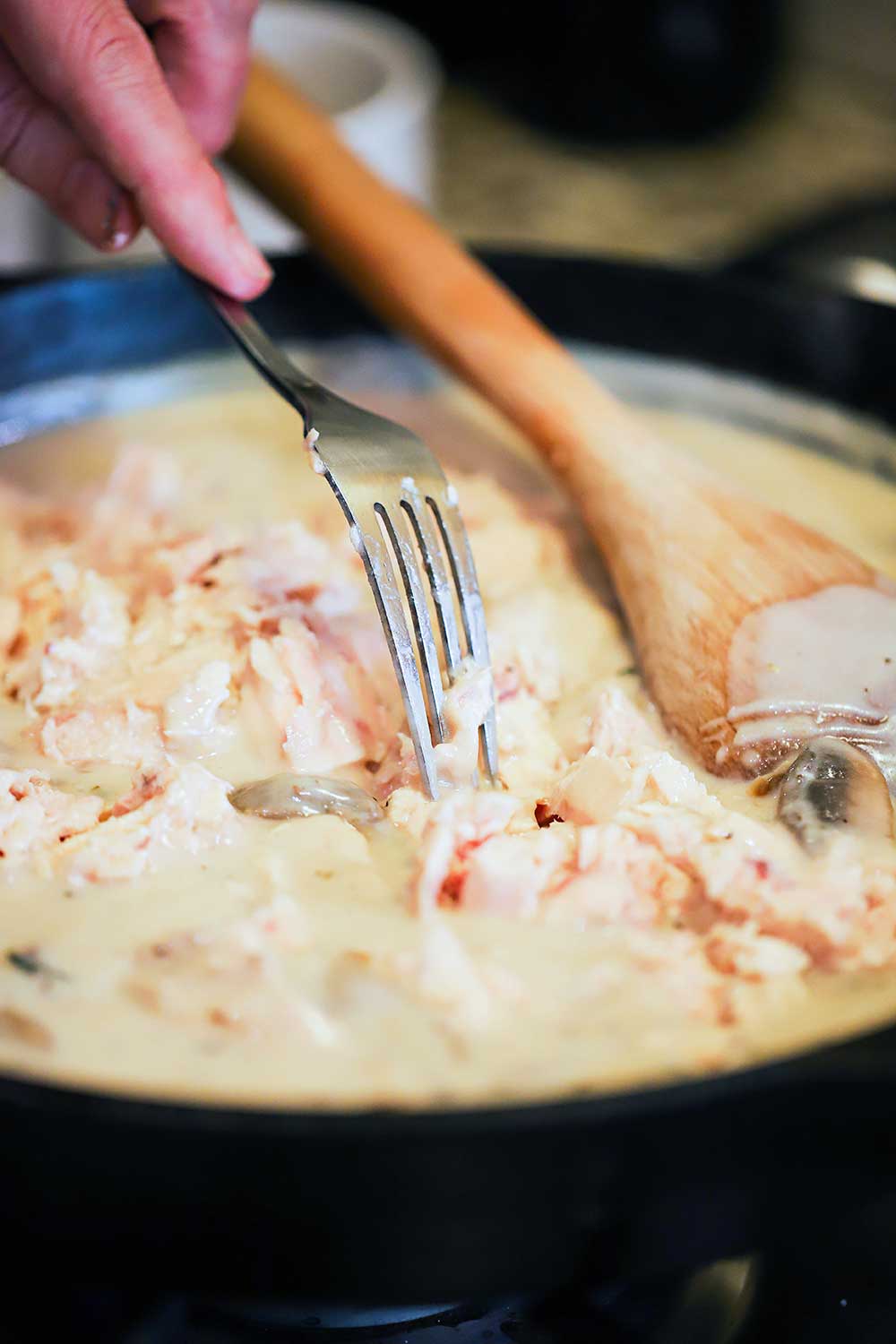 A person's hand using a fork to flake canned tuna into a cast-iron skillet filled with homemade cream of mushroom soup.