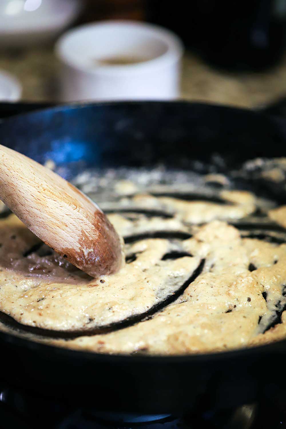 A wooden spoon stirring a blonde roux in a large black cast-iron skillet.