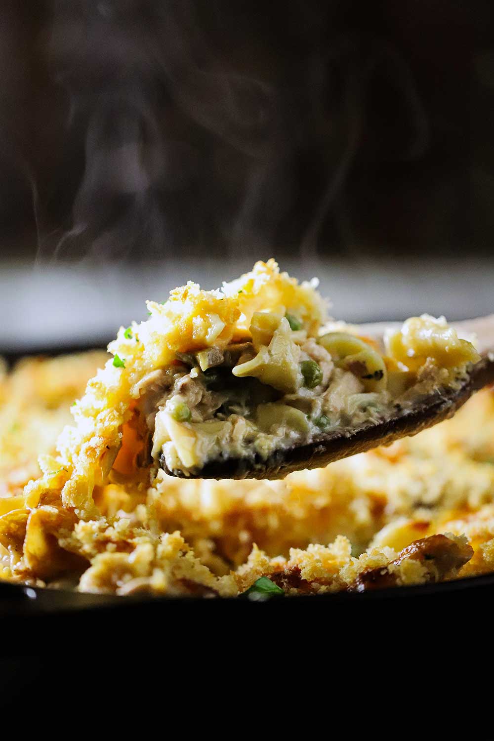 A large wooden spoon lifting a helping of cooked tuna casserole out of a black cast-iron skillet.