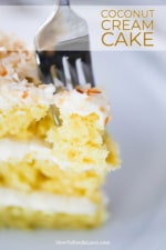 A fork inserted into the top of a piece of coconut cream cake standing upright on a white plate.