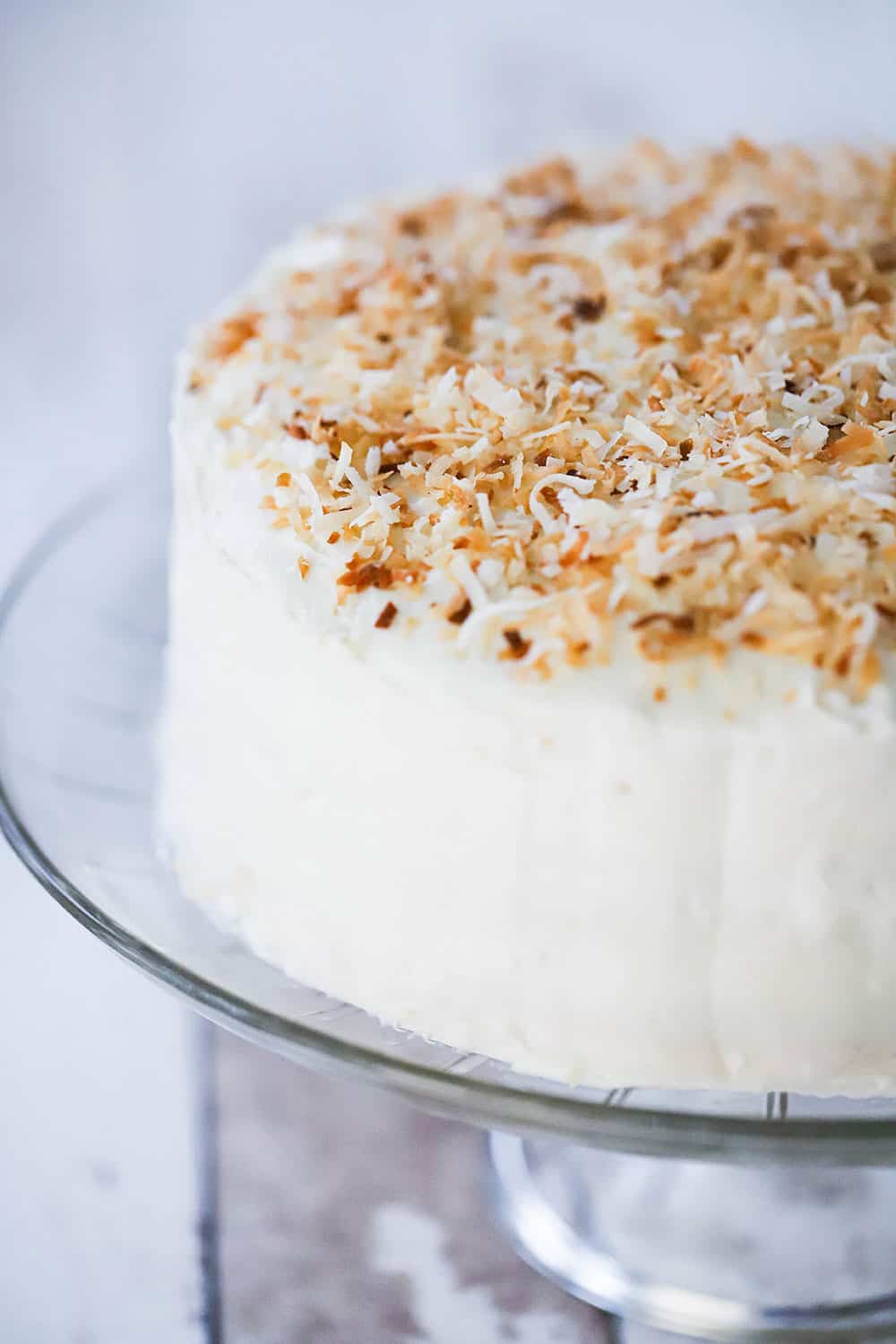 A coconut cream cake on a cake stand with toasted coconut flakes all over the top of the cake.