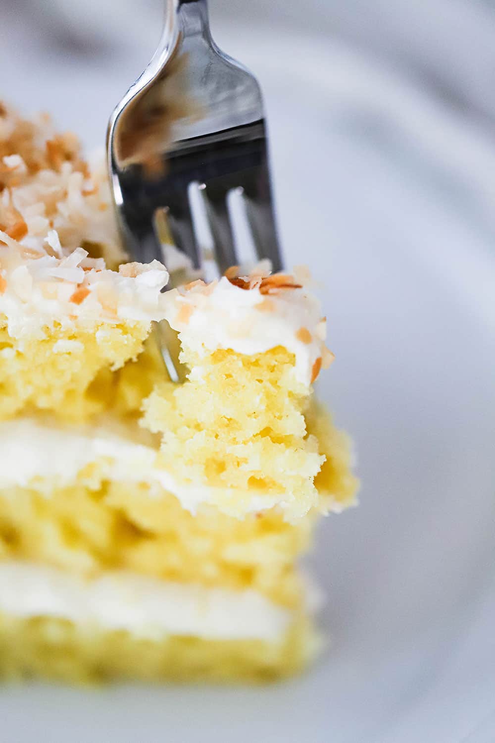 A fork inserted into the top of a piece of coconut cream cake standing upright on a white plate.
