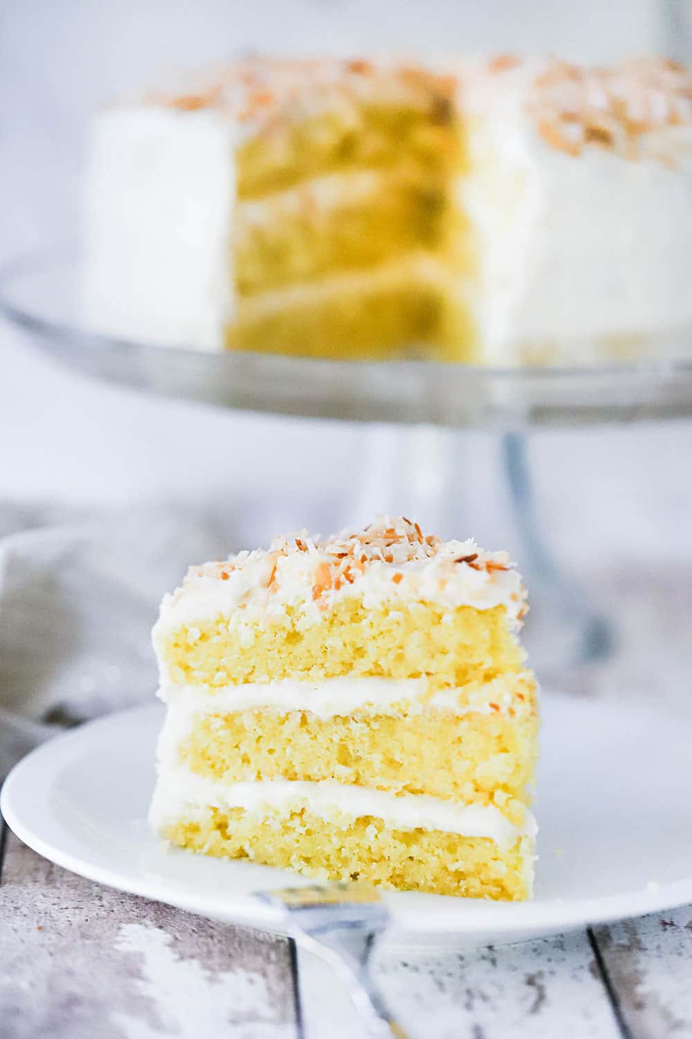A 3-layered slice of coconut cream cake standing upright on a plate in front of the cake on a cake stand.